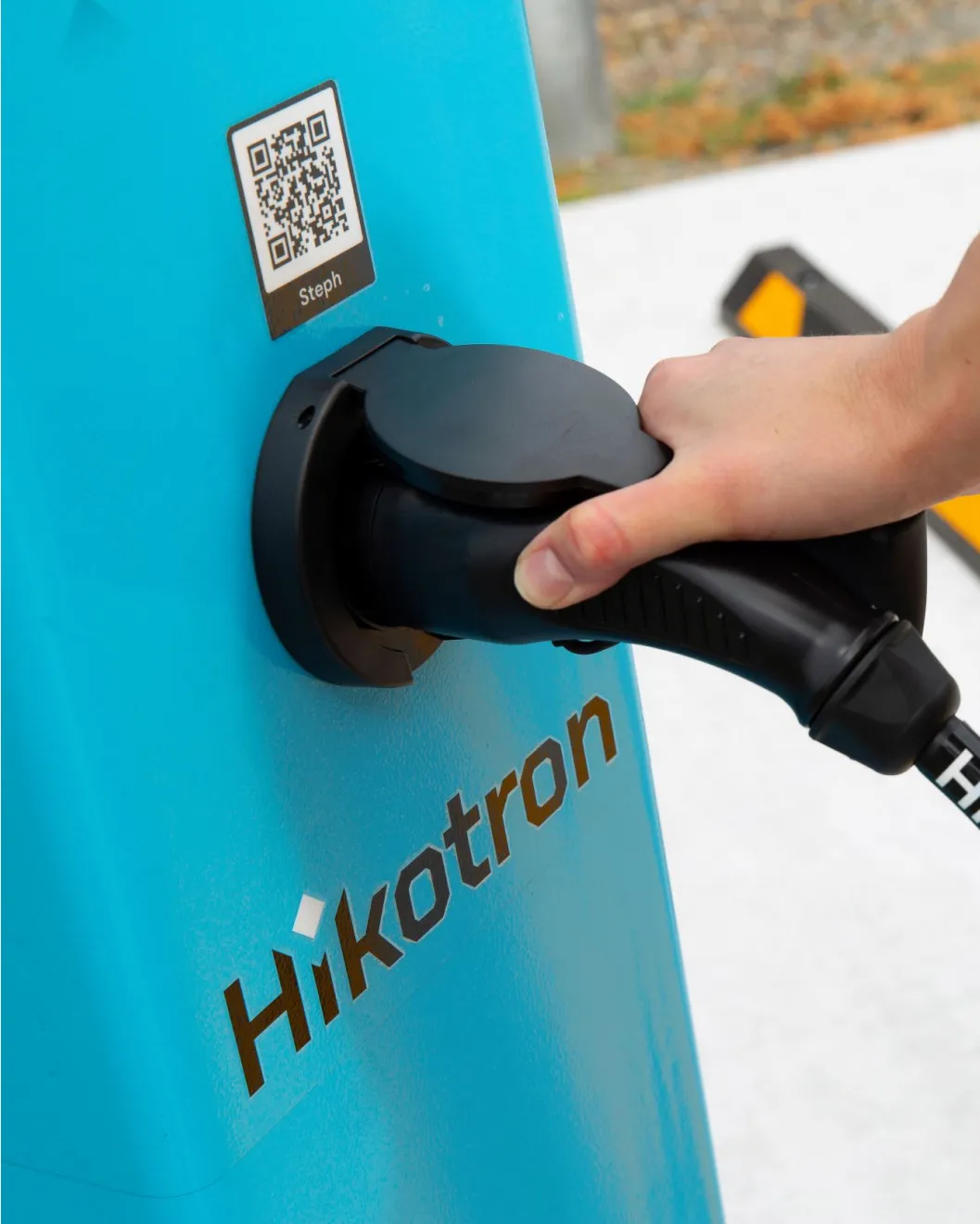 Hand plugging a Type 2 connector into a teal Hikotron charger with a QR code