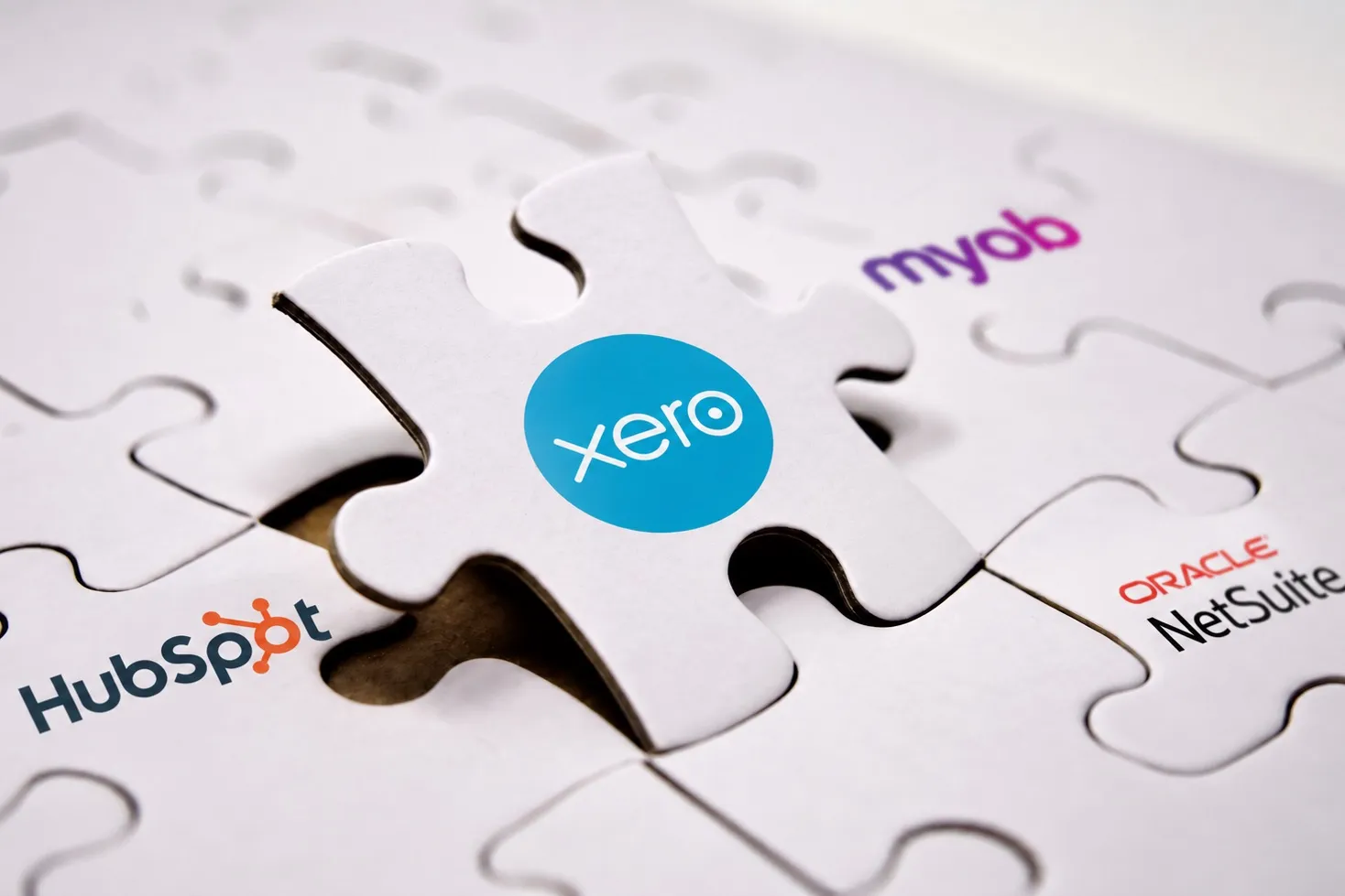 A jigsaw puzzle with a Xero branded piece being placed into a gap, surrounded by pieces branded MYOB, HubSpot and Oracle NetSuite.