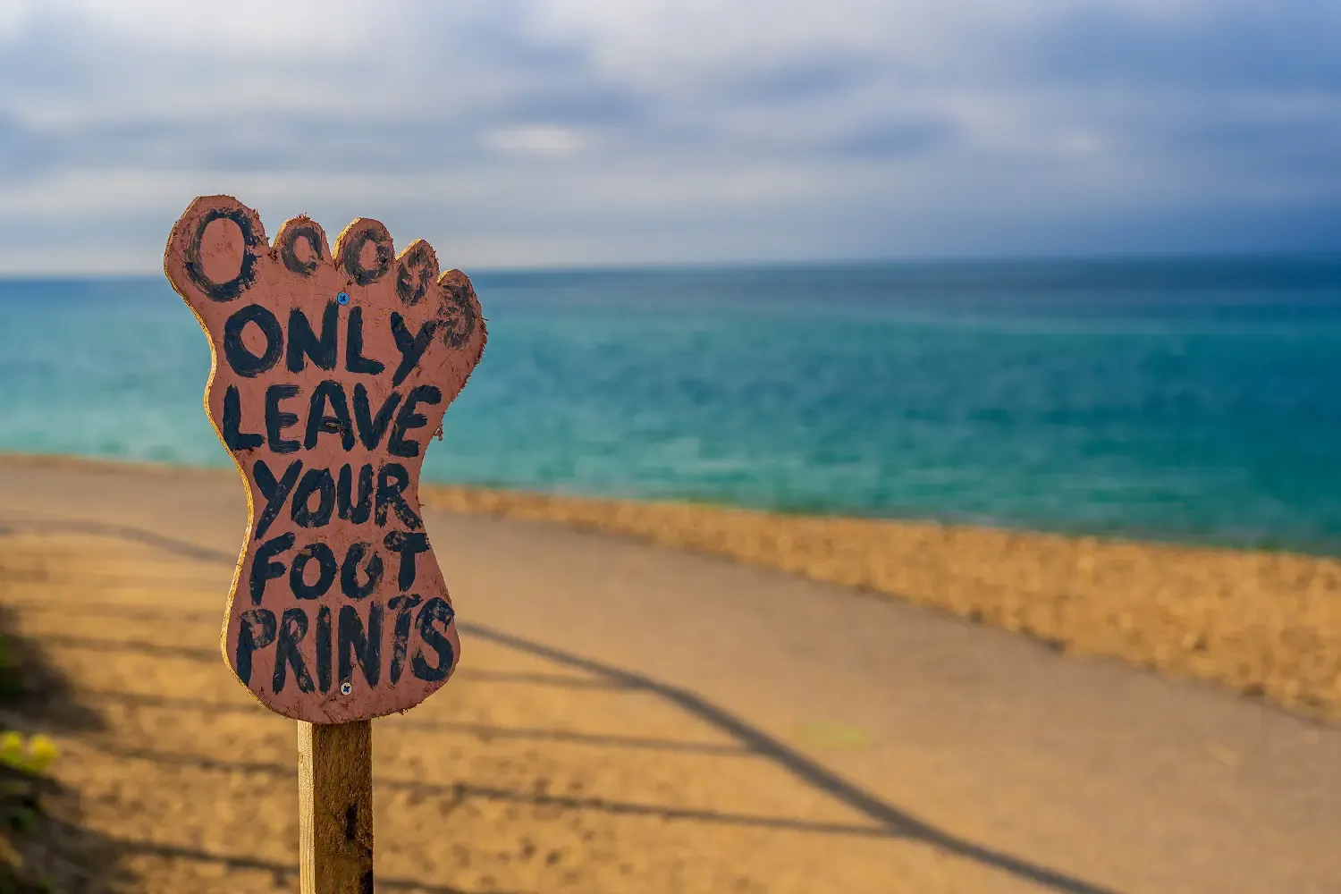 Hand-painted foot-shaped wooden beach sign reading "Only leave your footprints" beside the sea