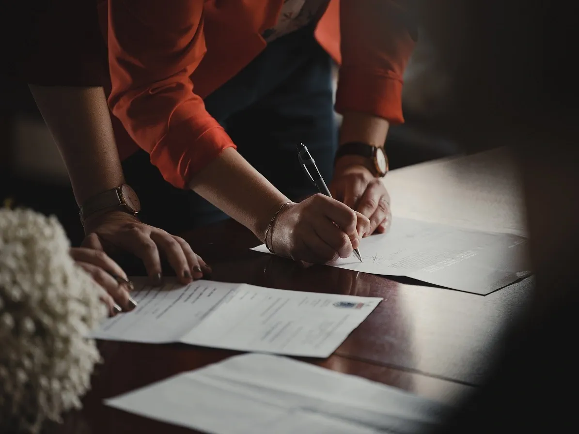 Hands signing paper contracts with a pen at a desk, representing slow manual paper-based quoting
