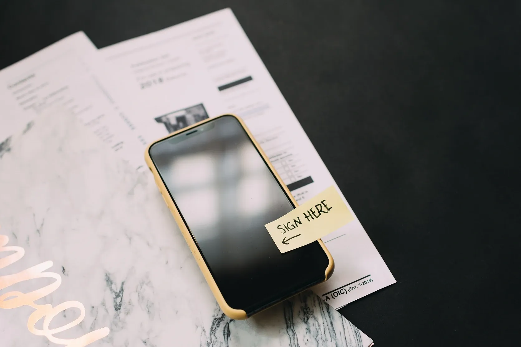 A smartphone resting on paper tax forms with a yellow Sign Here sticky note, contrasting paper and digital signing