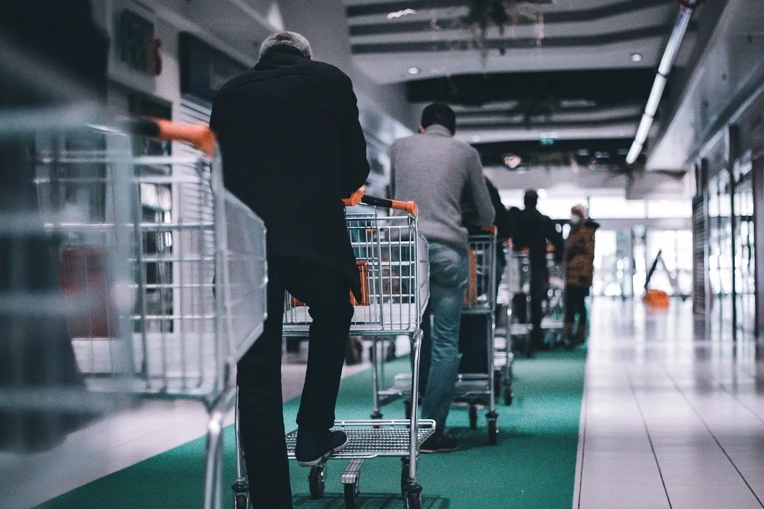 Shoppers with trolleys waiting single file in a store aisle, the physical queue virtual queuing replaces