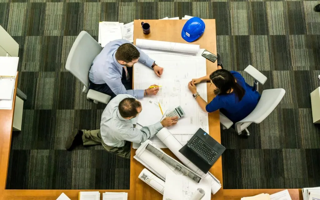 Overhead view of three professionals reviewing blueprints, a calculator and a hard hat while planning a build